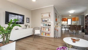 A modern living room with a flat-screen TV, a bookshelf, a dining area with a white table, and a kitchen in the background. - Family-friendly,air-conditioned, quiet location, garden, barbecue, pets welcome. (Porec)