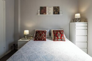 Een slaapkamer met een bed, twee kussens, een nachtkastje met een lamp, een dressoir en een kunstwerk aan de muur.