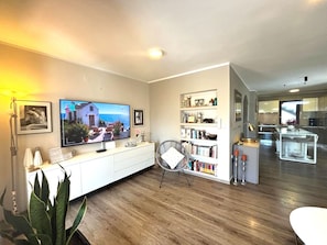 A modern living room with a flat-screen TV, a bookshelf, a dining area with a glass table, and a kitchen in the background.