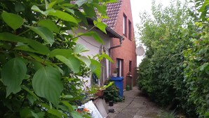 A narrow pathway leading to a house with a tiled roof and a brick wall. There are potted plants and a blue trash bin near the entrance. - Spacious u. child-friendly apartment (Oesterdeichstrich)