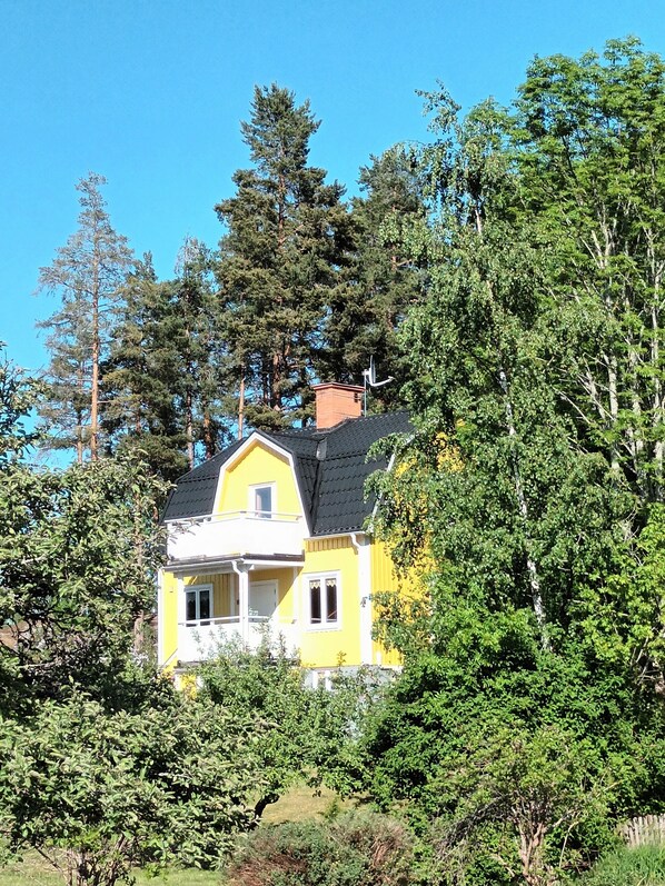 Une maison jaune, avec un toit noir, entourée d’arbres verdoyants et touffus.