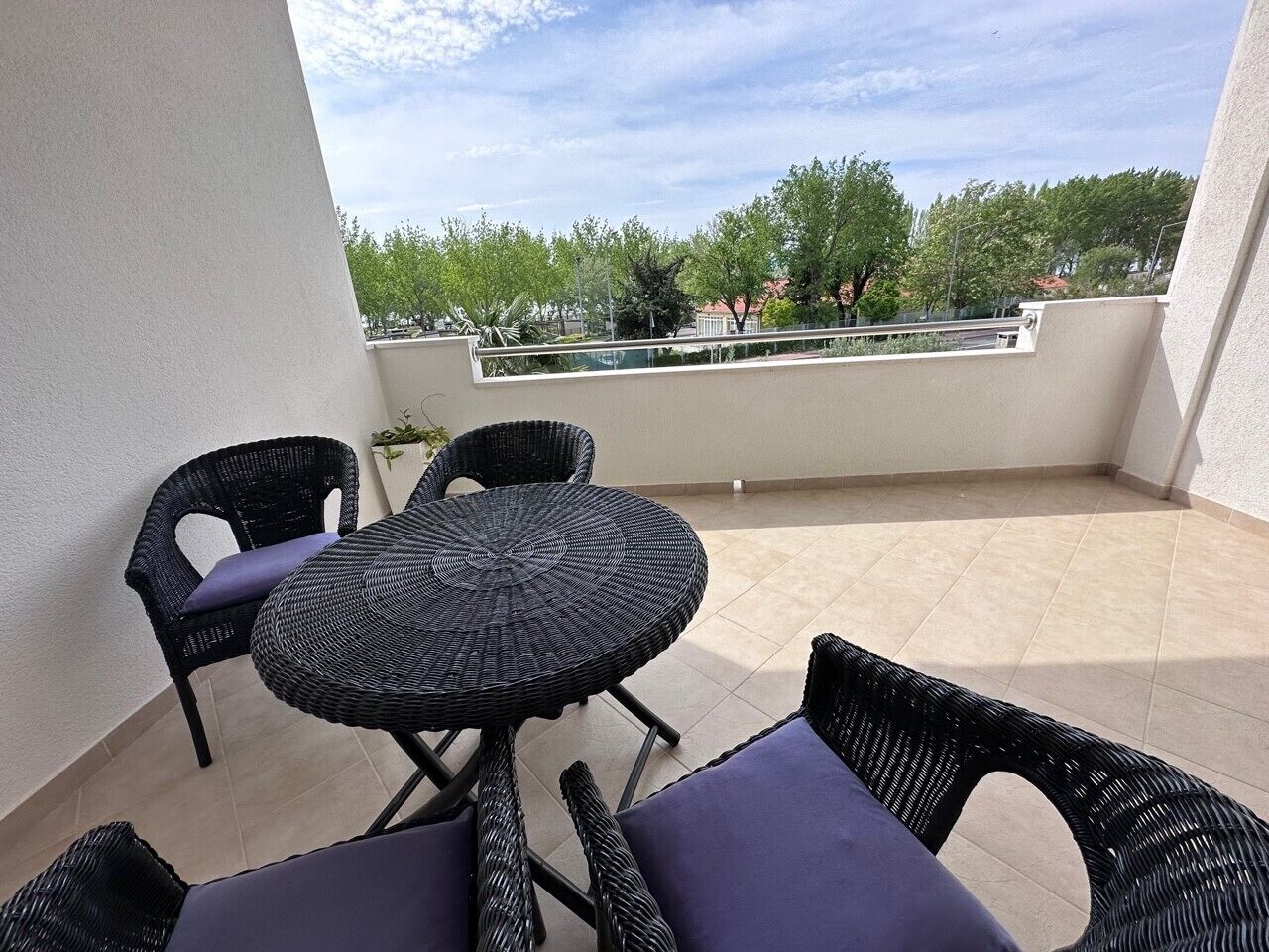 Un balcon agrémenté de meubles en osier, notamment une table ronde et deux chaises, d’où l’on peut admirer des arbres et des bâtiments en arrière-plan.