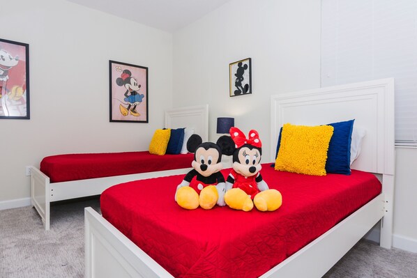 Une chambre avec deux lits, chacun recouvert d’une couverture rouge et doté d’une tête de lit blanche. On peut voir des peluches de Mickey et de Minnie Mouse sur les lits.