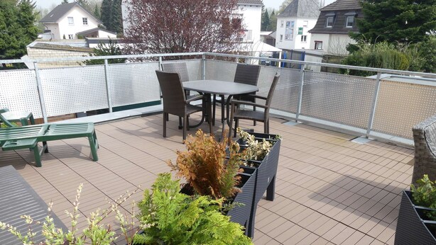 A rooftop terrace with a table and chairs, surrounded by potted plants and a metal fence.