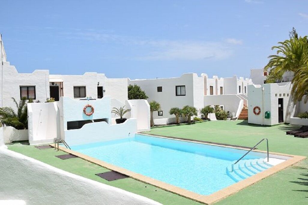 Un área de piscina con una piscina azul cristalina, rodeada de edificios blancos con balcones y pequeños árboles.