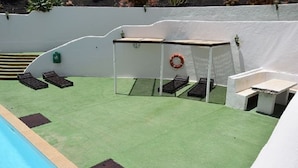 Zona de piscina con césped artificial verde, una pared blanca y una pequeña mesa con una silla.