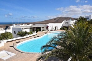 Une piscine entourée de bâtiments blancs, avec vue sur la mer et les collines en arrière-plan.