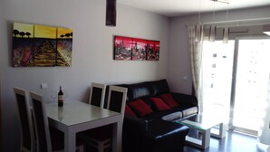 A modern living room with a dining area, featuring a sofa, a glass coffee table, and a dining table with chairs. There are two paintings on the wall, a cityscape and a landscape. - OFFERS FOR LAST DAYS!!, LUXURY ATTIC CARIHUELA,  2 GARAGES, POOL, INT. CHANNELS (Torremolinos)