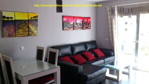 A modern living room with a black leather sofa, red cushions, a glass coffee table, and two white chairs. There are two paintings on the wall, one depicting a sunset and the other a cityscape. - OFFERS FOR LAST DAYS!!, LUXURY ATTIC CARIHUELA,  2 GARAGES, POOL, INT. CHANNELS (Torremolinos)