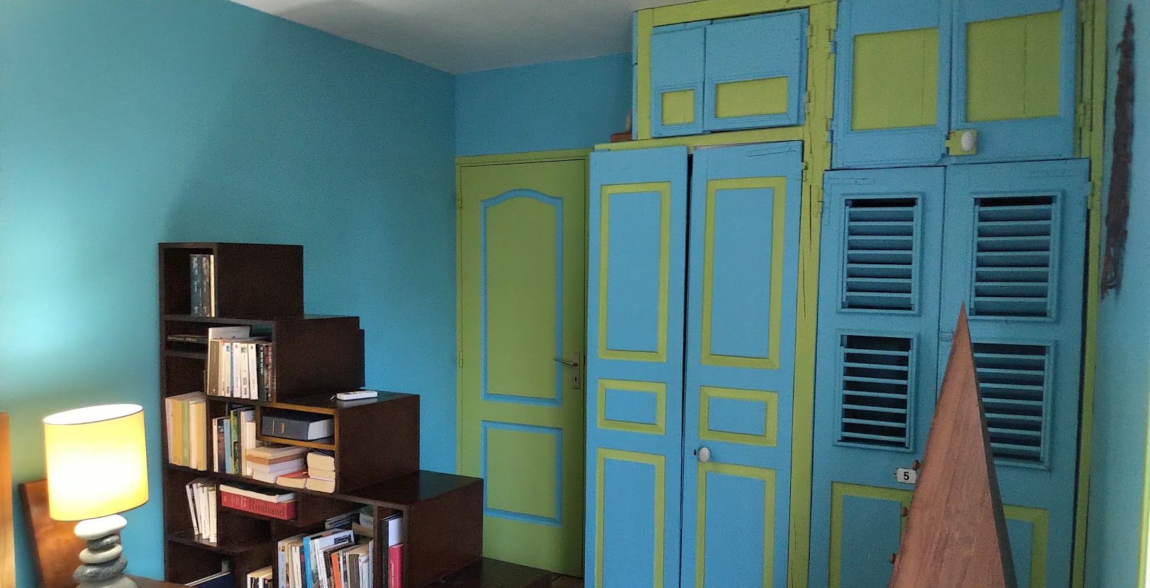 Une pièce avec une bibliothèque, une lampe et une porte ornée de panneaux bleus et jaunes.