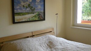 A bedroom with a bed, a painting of a landscape, a window with a view of greenery, and a wall-mounted light fixture.