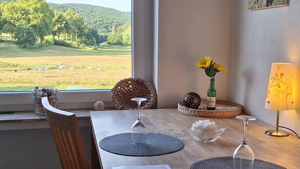Een eettafel met placemats, een wijnglas en een lamp. Door het raam is een groen landschap zichtbaar.