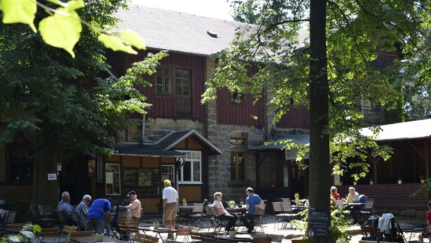 An outdoor café with tables and chairs, surrounded by trees and a stone building with wooden shutters.