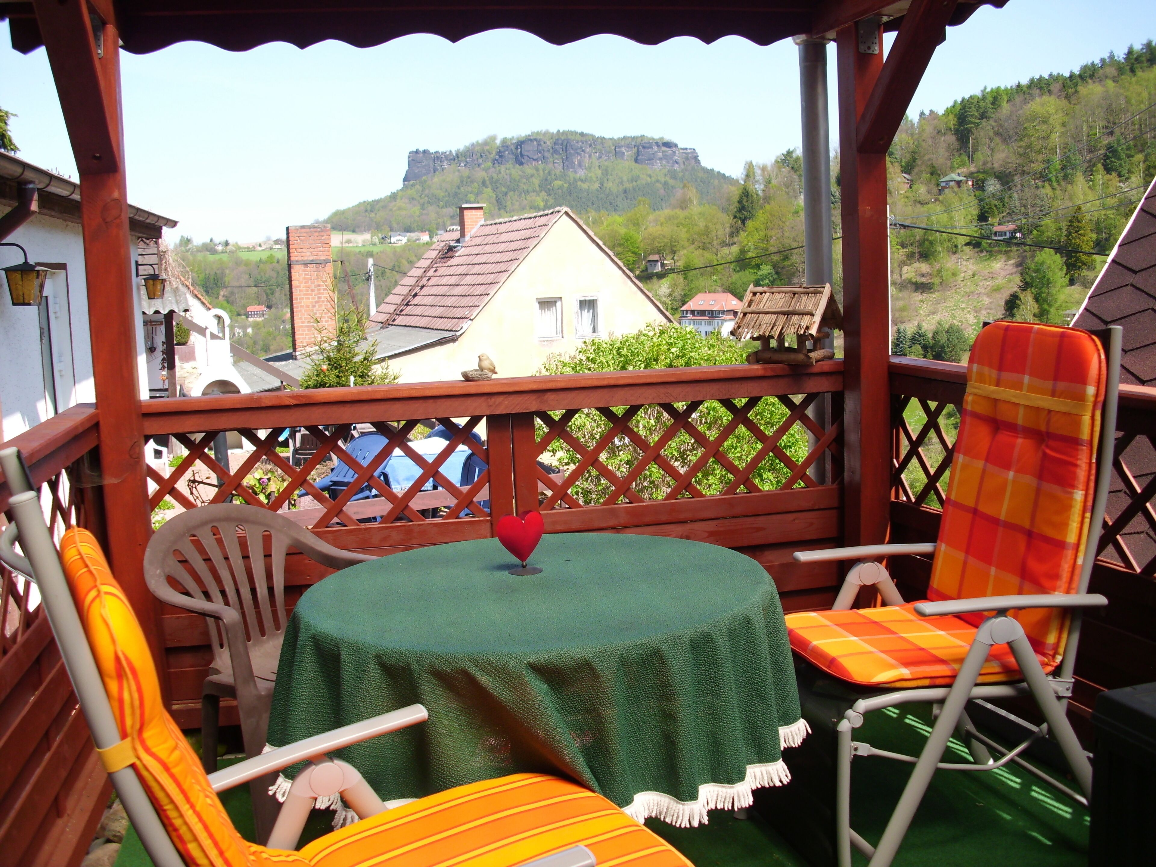 A covered outdoor seating area with a table and chairs, overlooking a village and a mountain.