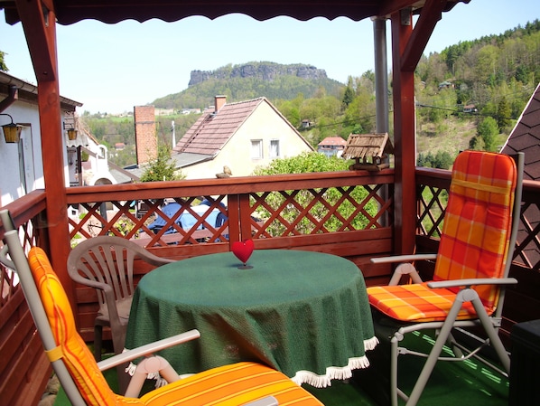 A covered outdoor seating area with a table and chairs, overlooking a village and a mountain.
