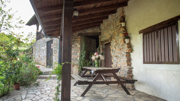 Un pati de pedra amb taula i bancs de picnic de fusta, envoltat de vegetació i un edifici de pedra amb bigues i persianes de fusta.