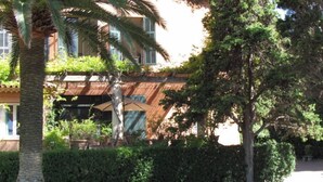 Een gebouw met een traditionele architectuur, herkenbaar aan het pannendak en het balkon. Een verzorgd gazon bevindt zich voor het gebouw, en er zijn verschillende bomen en struiken rondom het perceel.