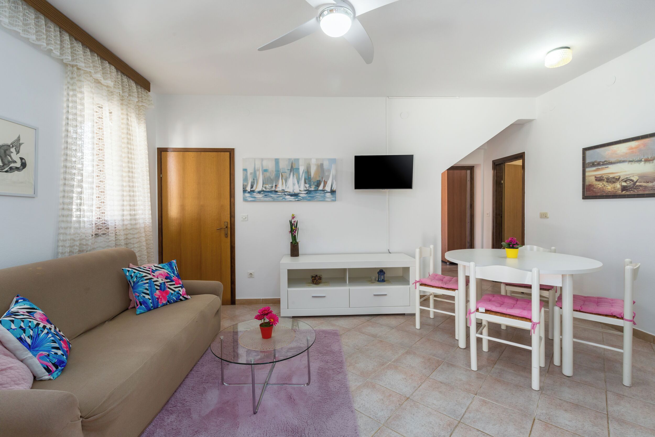 A living room with a sofa, coffee table, and a dining table with chairs. There is a TV mounted on the wall, a cabinet, and a door.