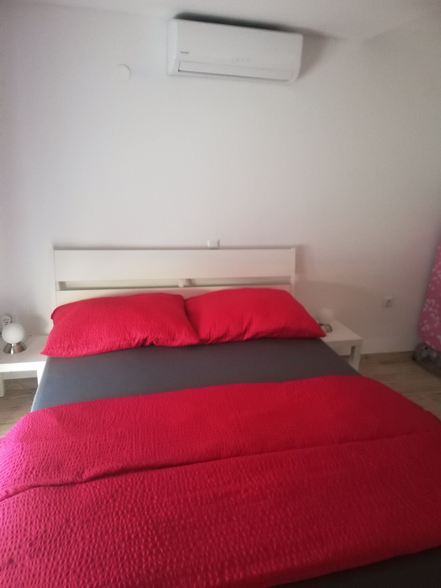 A bed with red and grey bedding, a white headboard, and a white nightstand with a lamp.