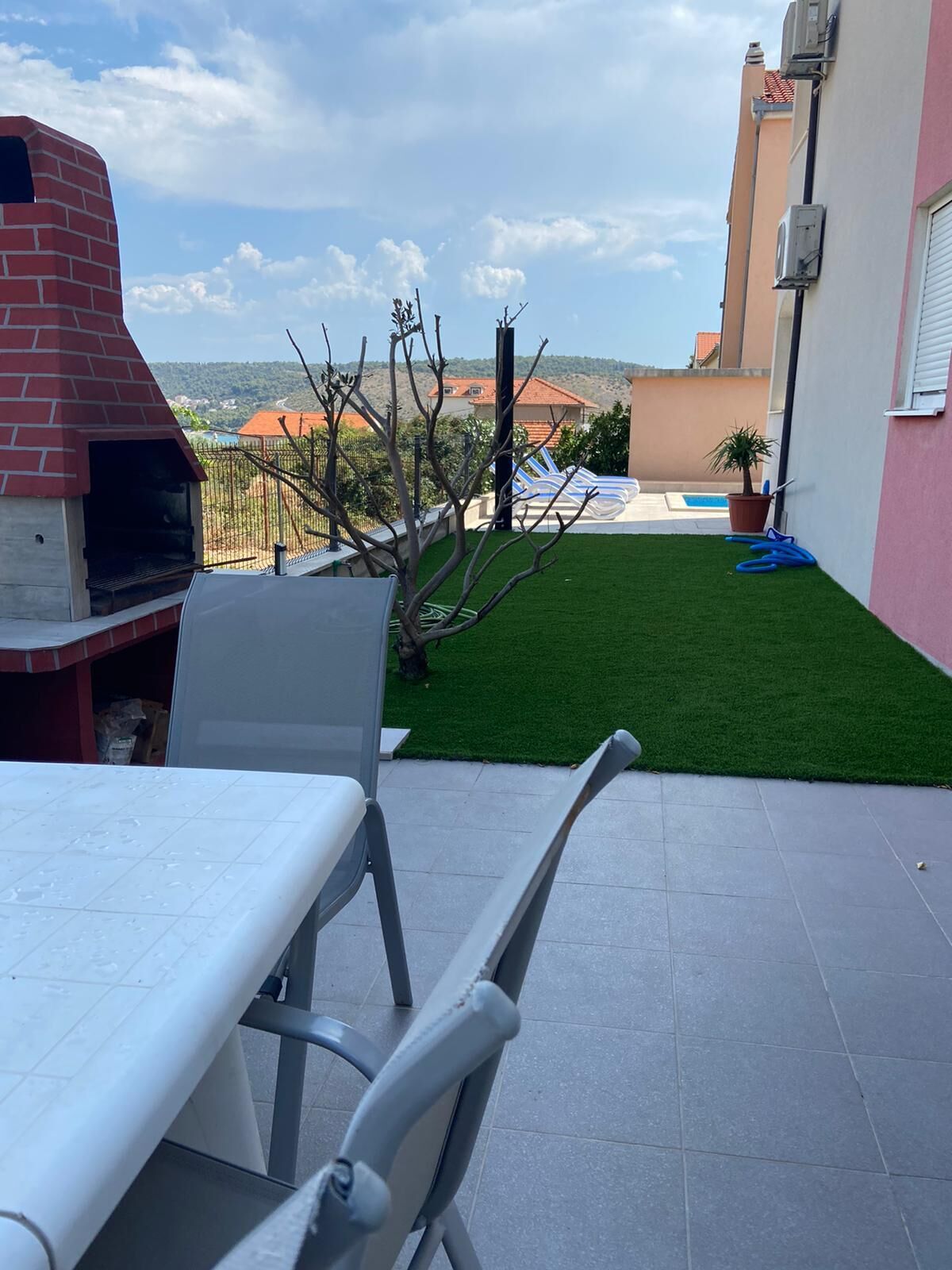 Un patio con forno in mattoni, un tavolo e una sedia. Sullo sfondo, una piscina e vista su case e colline.