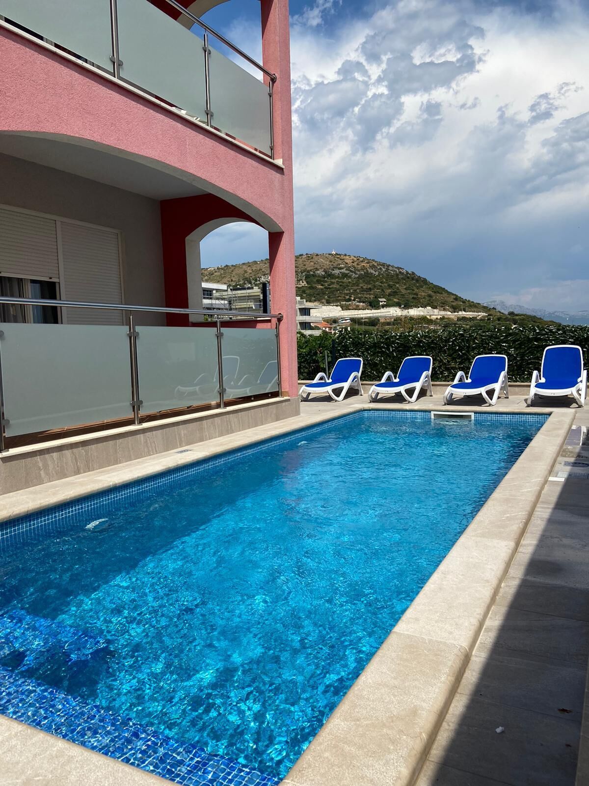 Ein Swimmingpool mit blauen Fliesen, umgeben von Liegestühlen und einem Gebäude mit rosafarbenem Mauerwerk.
