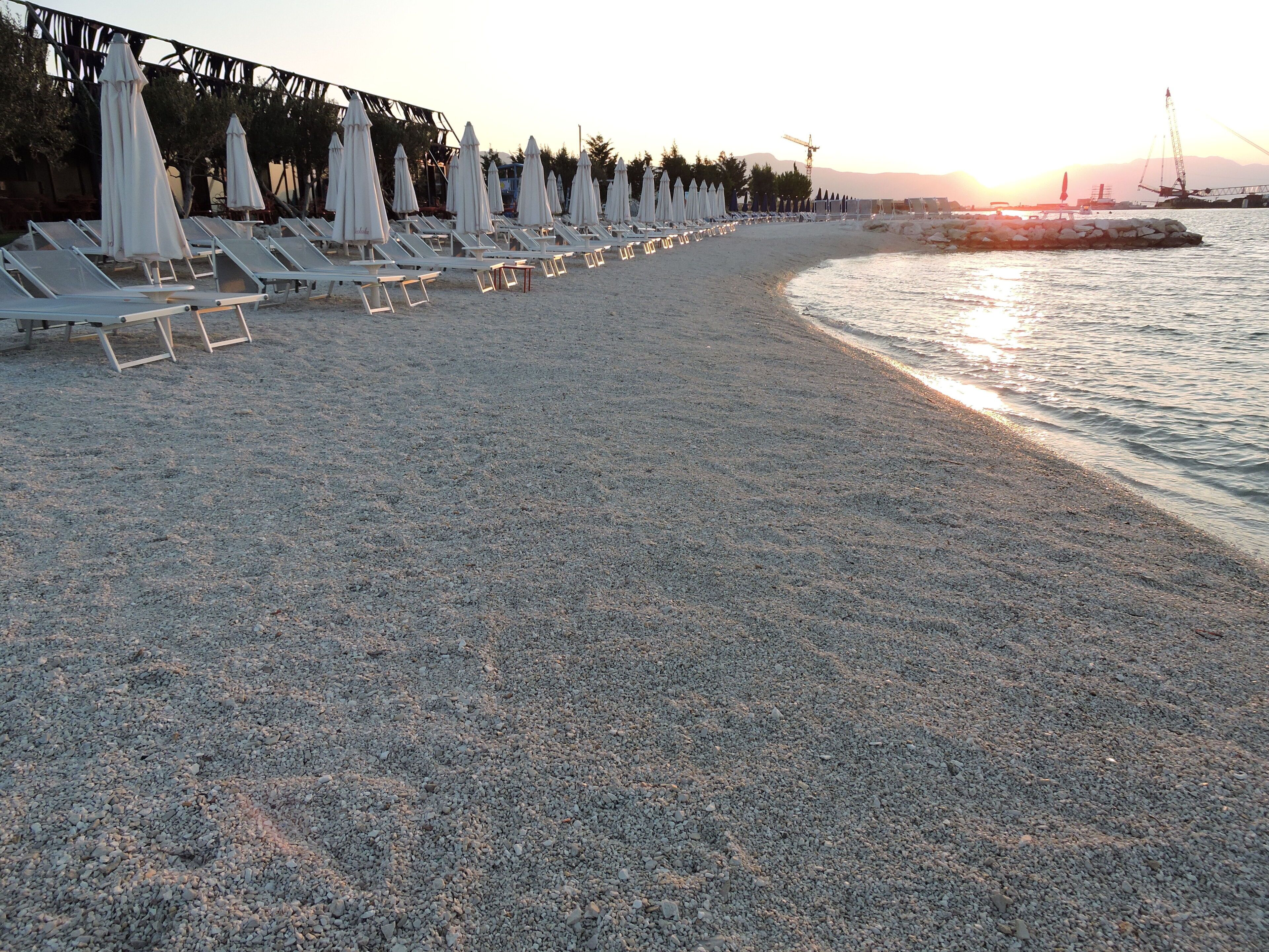 Una spiaggia con ciottoli, file di lettini e ombrelloni, un molo sullo sfondo e un tramonto sull'acqua.