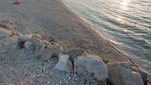 A pebbled beach with lounge chairs and umbrellas, a rocky foreground, and a calm sea at sunset.