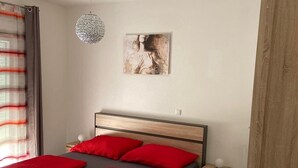 A bedroom with a wooden bed, red pillows, a bedside table, a hanging light fixture, and a painting on the wall.