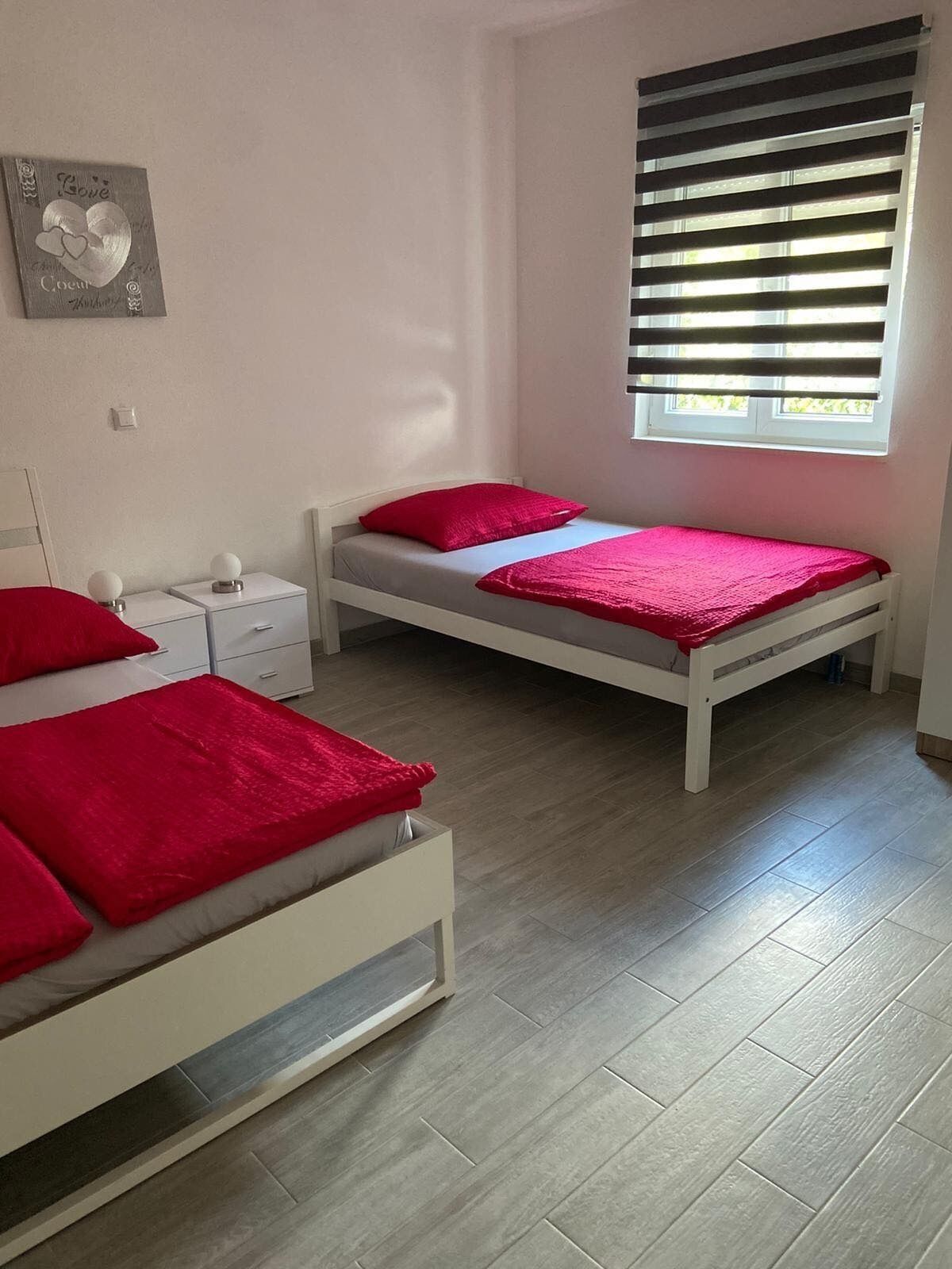 Une chambre avec deux lits simples, une table de chevet, une petite table et une fenêtre avec des stores.