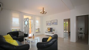Een moderne woonkamer met zwarte leren banken, een witte salontafel en een televisie op een witte dressoir. Twee ingelijste foto's hangen aan de muur en daarboven een kroonluchter.