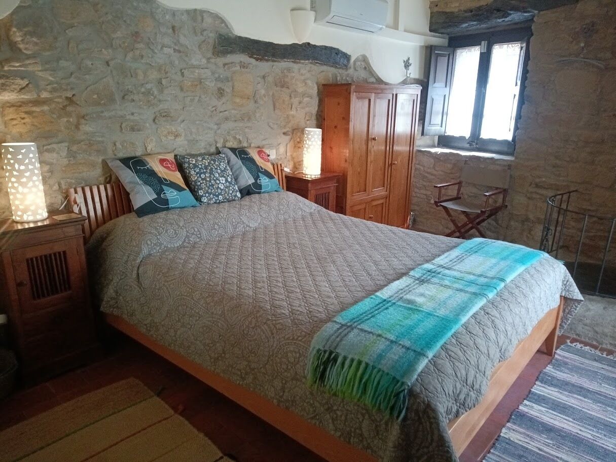 Quarto com parede de pedra, cama de madeira, mesa de cabeceira com abajur e janela com cortinas.