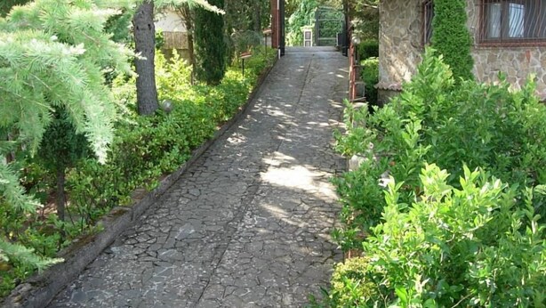 Un chemin de pierre mène à un bâtiment en pierre surmonté d’un toit en tuiles, le tout étant entouré d’une végétation luxuriante et d’arbres.