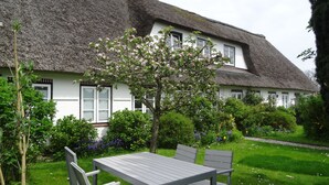 A thatched-roof house with a white exterior, a garden with a table and chairs, and blooming trees. - Holidays by the sea in a historic thatched house: WiFi, 2 Smart TVs, sauna from 2022 (Pommerby)
