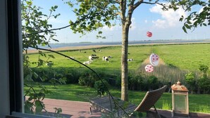 A view from a window overlooking a grassy field with cows, a tree, and a wind turbine in the distance. - Vacation apartment in the Rosenhaus am See (Dagebüll)