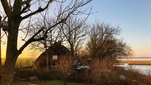 A rustic wooden cabin with a thatched roof, surrounded by a grassy field and a body of water. - Vacation apartment in the Rosenhaus am See (Dagebüll)
