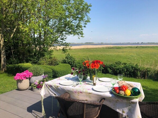An outdoor dining table set with plates, cutlery, and a bowl of fruit, surrounded by greenery and a view of wind turbines.