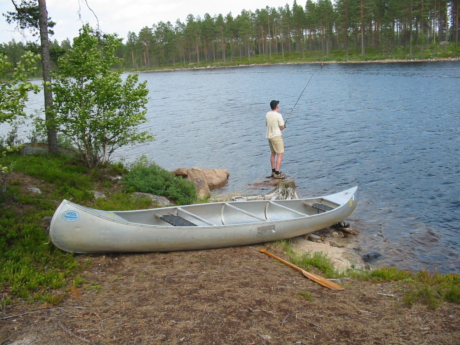 Una canoa en la orilla con una persona pescando, rodeada de árboles y un lago tranquilo.