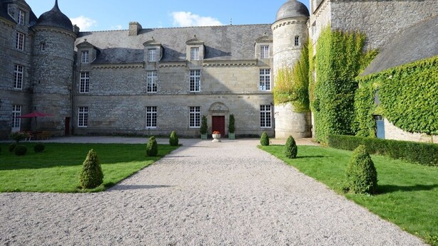 Un bâtiment historique en pierre, doté d’une cour intérieure, d’une porte rouge et d’un chemin de gravier menant à l’entrée.