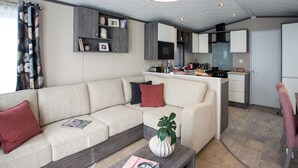 A modern living room with a sofa, a coffee table with a magazine, and a kitchen area in the background. - 3 Bed Caravan, South Devon Coast nr Plymouth, 200m to Beach + Indoor Heated Pool (Nr Plymouth)