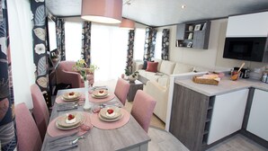A modern dining area with a wooden table set for four, surrounded by pink chairs. A large pendant light hangs above the table, and a kitchen area is visible to the right with a microwave and a television. - 3 Bed Caravan, South Devon Coast nr Plymouth, 200m to Beach + Indoor Heated Pool (Nr Plymouth)