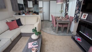 A compact living space with a sofa, coffee table, dining table, and chairs. A kitchen area is visible in the background. - 3 Bed Caravan, South Devon Coast nr Plymouth, 200m to Beach + Indoor Heated Pool (Nr Plymouth)