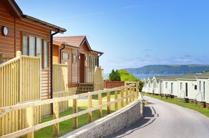 A row of wooden cabins with a stone wall and wooden fence in the foreground, and a clear view of the sea in the background. - 3 Bed Caravan, South Devon Coast nr Plymouth, 200m to Beach + Indoor Heated Pool (Nr Plymouth)