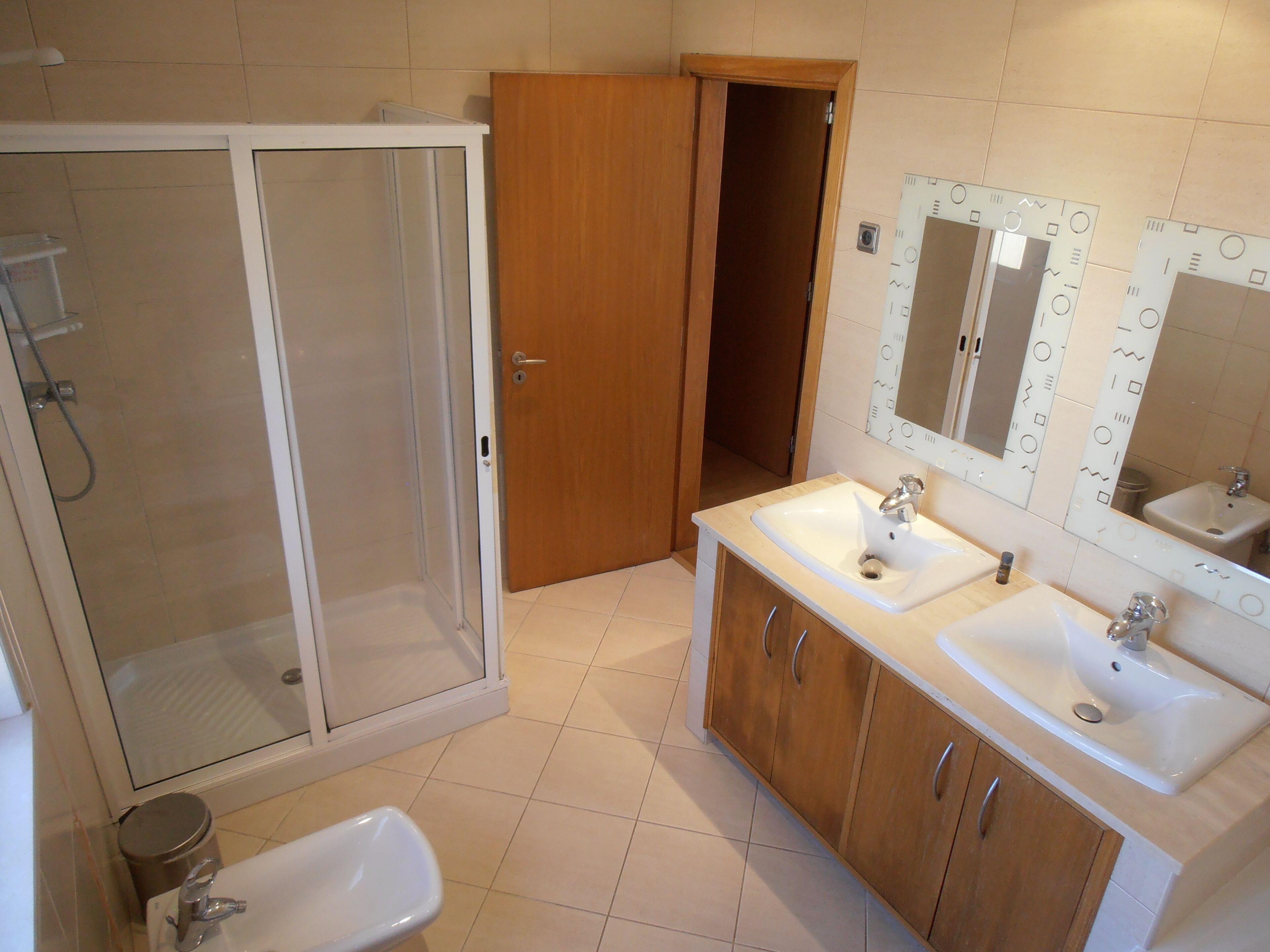 Ein Badezimmer mit Dusche, zwei Waschbecken, Holzschränken und einem Spiegel mit dekorativen Mustern.