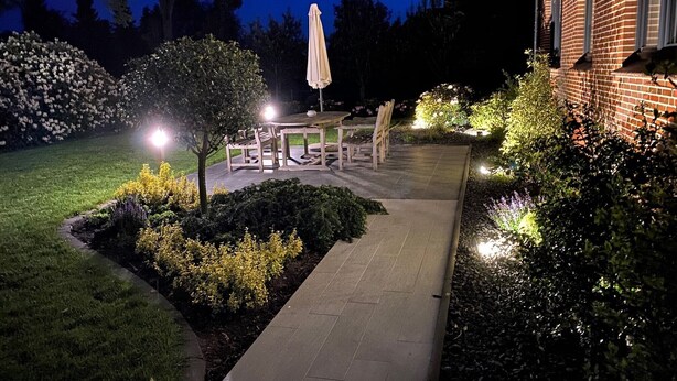 A patio area with a table and chairs, surrounded by well-lit garden beds and a pathway leading to a building. - 5-star luxury apartment Albatros directly on the golf course with its own garage and terrace (Wiesmoor)
