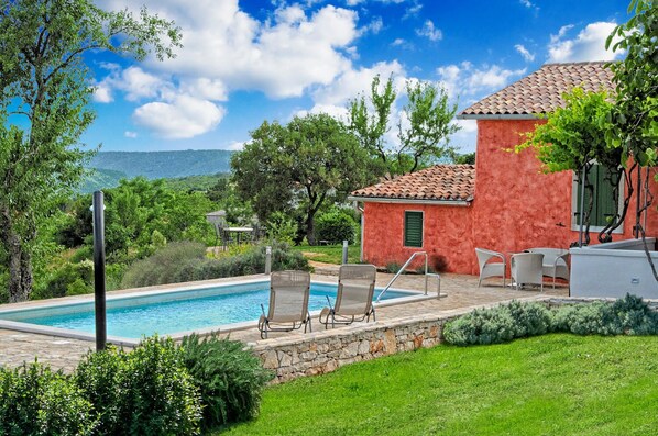 A swimming pool with lounge chairs, a stone wall, and a red building with a tiled roof.