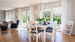 A spacious dining area with a white table and chairs, large windows with white curtains, and a view of greenery outside.
