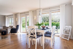 Un comedor amplio con una mesa y sillas blancas, ventanas grandes con cortinas blancas y vistas a zonas verdes.