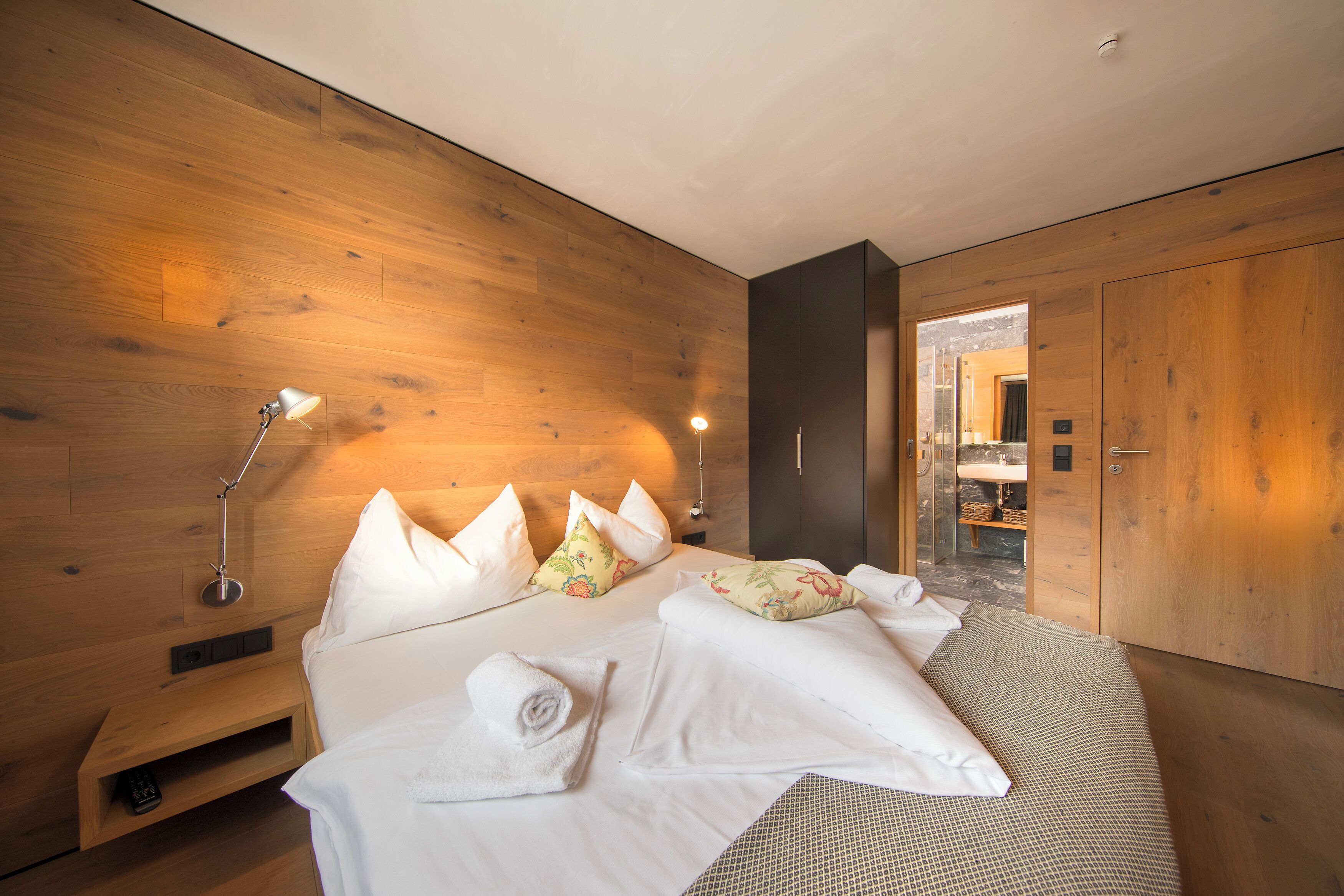 Ett modernt hotellrum med en väggpanel i trä, en säng med vita sängkläder, en sänglampa och utsikt in i badrummet.