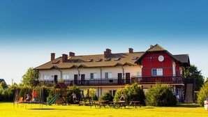 A two-story building with a red and white facade, a balcony, and a playground area with a slide and swings. - Family-friendly, green area, barbecue area, surrounded by pastures, nature park  (Rowy)