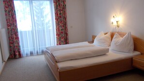 Cama doble con ropa de cama blanca, mesita de noche, lámpara de pared y ventana con cortinas florales.